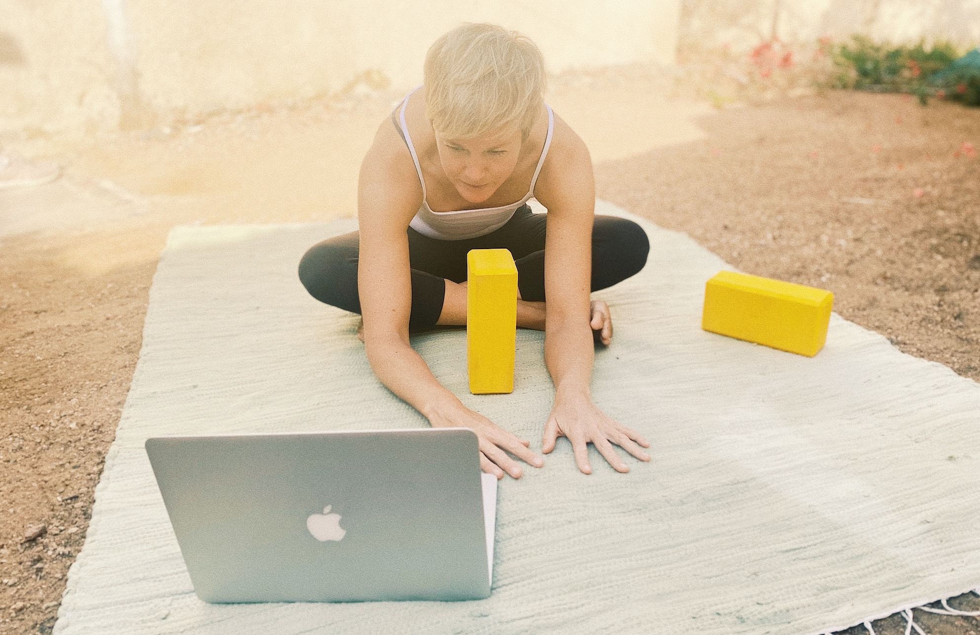 Kostenfreie Downloads | Nina Raem draußen auf einer Matte mit Yogaklötzen vor dem Laptop sitzend und auf den Bildschirm blickend